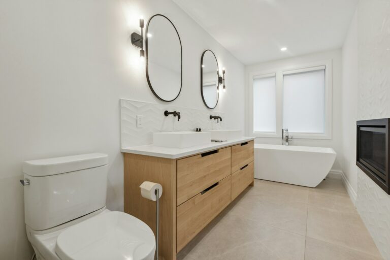 Moderne badkamer met toilet, dubbele wastafel met houten kasten, twee ovale spiegels, wandlampen, een vrijstaand bad en een open haard. Witte muren en grote tegels maken het ontwerp compleet.