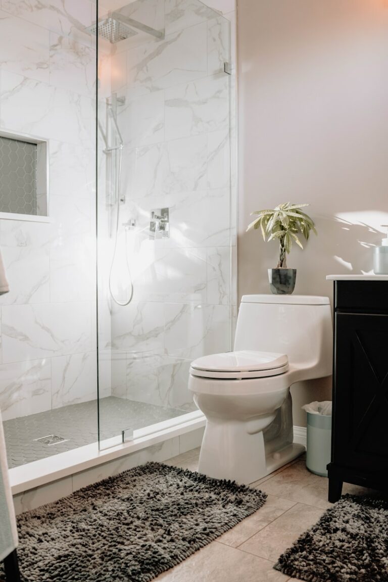 Moderne badkamer met een glazen douche, witte marmeren tegels, een wit toilet, een klein potplantje op het reservoir van het toilet en donkergrijze tapijten op de vloer.