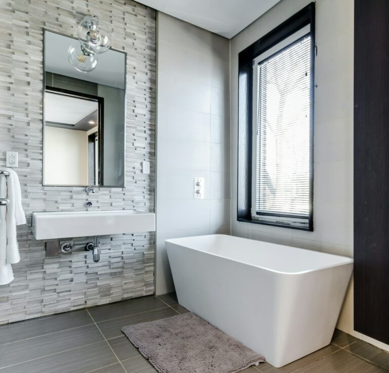 Moderne badkamer met een aan de muur gemonteerde wastafel, rechthoekige spiegel, groot wit bad, vloermat en raam met jaloezieën. Grijze getextureerde wandtegels en donkere vloertegels maken het decor compleet.