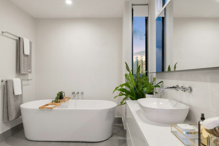 Moderne badkamer met een vrijstaand wit bad, dubbele wastafel op een strak aanrechtblad, handdoekenrekken en een grote plant naast een raam.