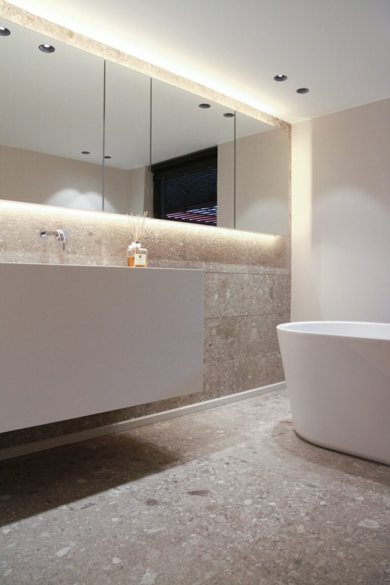 Moderne badkamer met een grote spiegel, zwevend wastafelmeubel, strak aanrechtblad, rechthoekige wastafel, vrijstaand bad en subtiele verlichting. Neutrale tinten en stenen vloeren creëren een minimalistische sfeer.