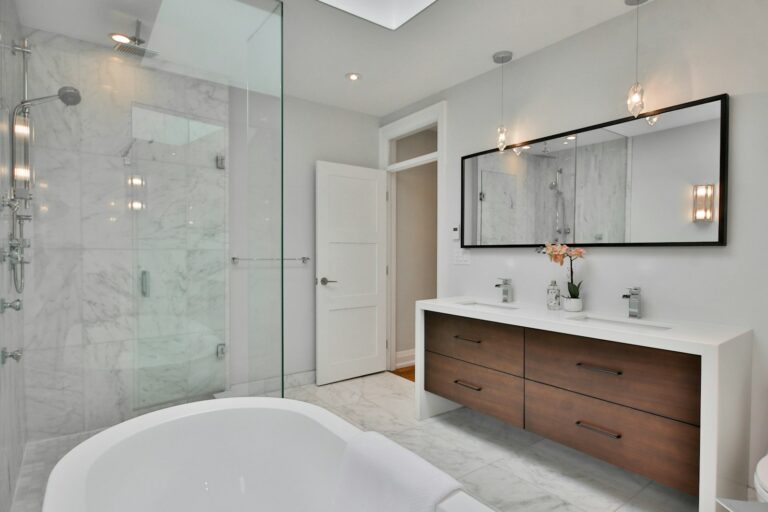 Moderne badkamer met marmeren tegels, glazen douche, ligbad, dubbele wastafel met houten lades, grote spiegel en hanglampen.