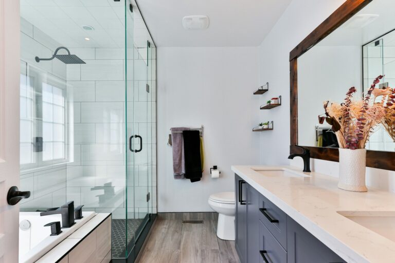 Moderne badkamer met glazen douche, bad en dubbele wastafel. Met een grote spiegel, witte muren en houten vloer.