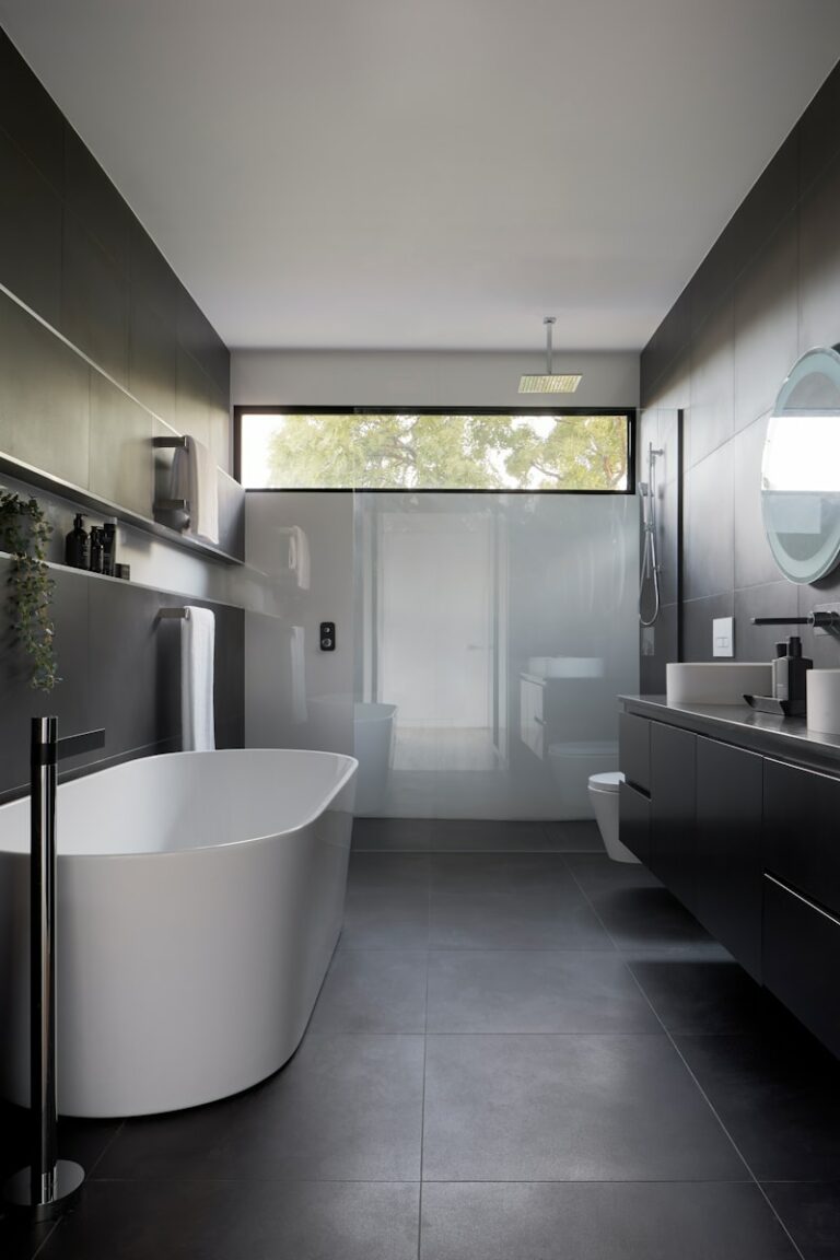 Moderne badkamer met een vrijstaand bad, zwevende wastafel, ronde spiegel en een raam met natuurlijk licht. Donkere tegels bedekken de muren en vloer, wat een strak uiterlijk creëert.