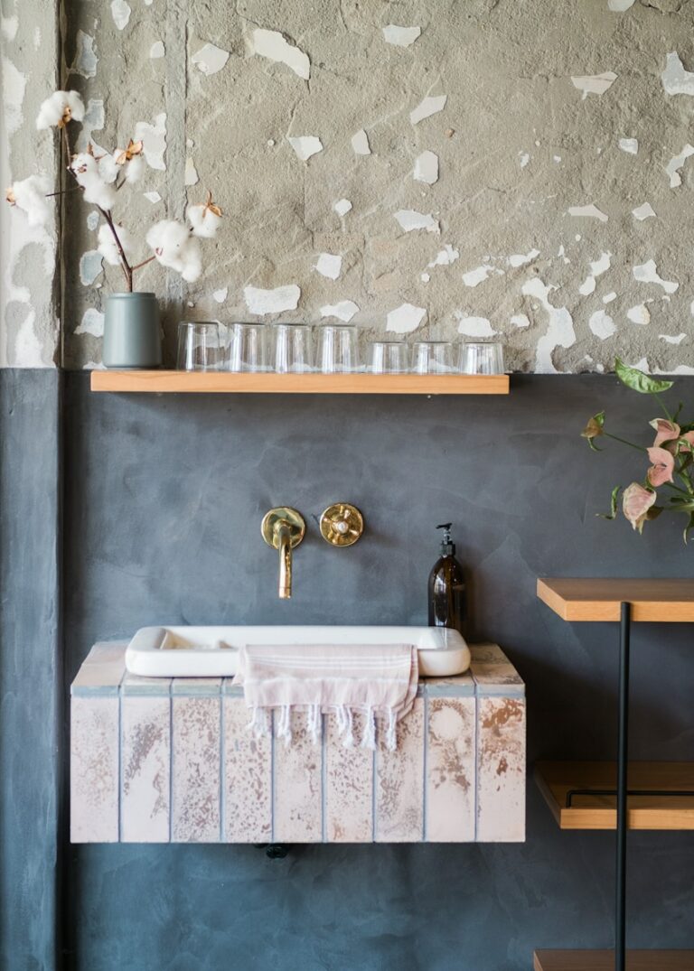 Een rustieke muur met een kleine gootsteen, een koperen kraan, een zwarte zeepdispenser en een handdoek. Bovenop een plank met glazen en een vaas met katoenen takken. Op een andere plank staat een vaas met bloemen.