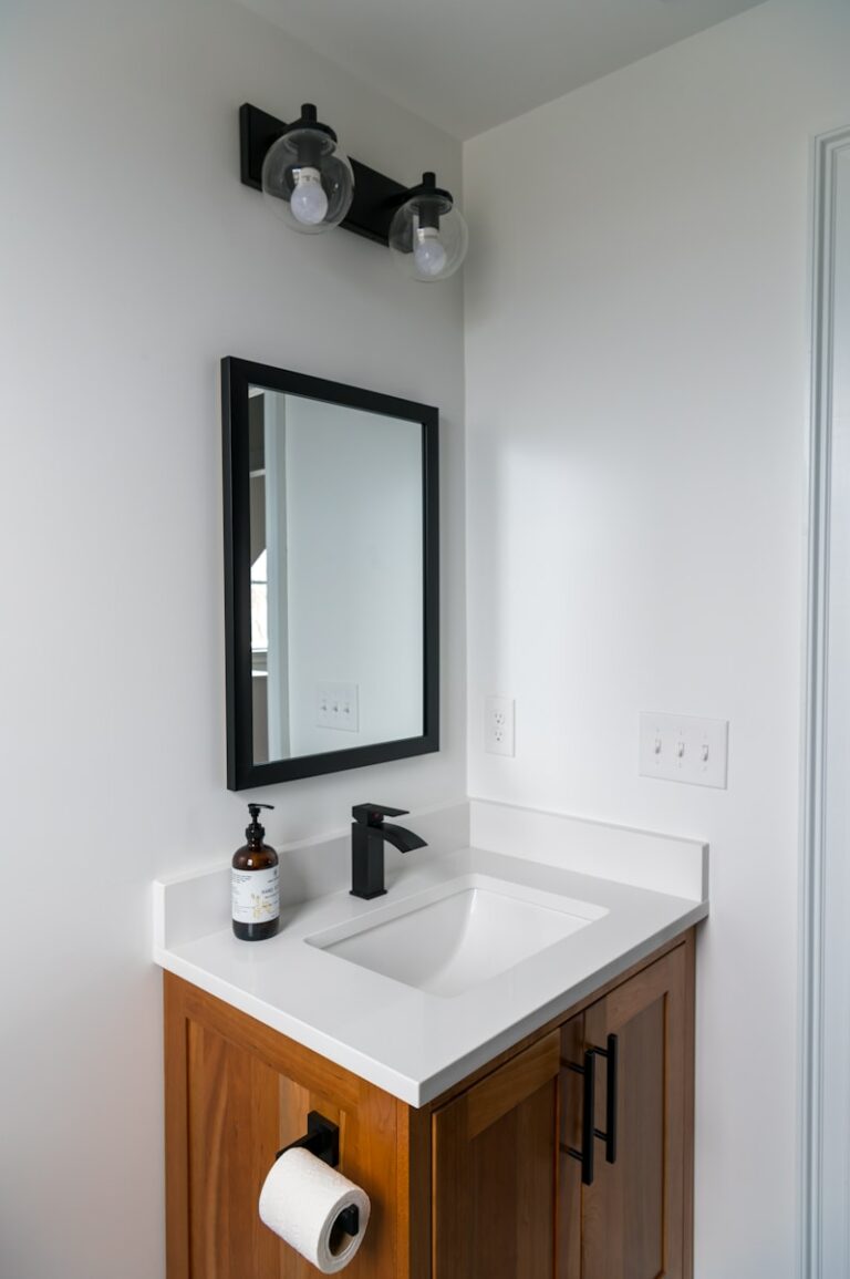 Badkamerhoek met een houten wastafel, wit aanrecht, zwarte kraan, aan de muur gemonteerde spiegel, zeepdispenser en een lamp met twee lampen erboven. Toiletpapierhouder bevestigd aan de wastafel.