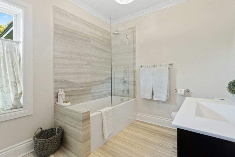 Moderne badkamer met een glazen badkuip, lichte houten accenten, witte handdoeken op een rek, een mand en een wastafel. Lichtbeige muren en gordijnen met patroon op een raam.