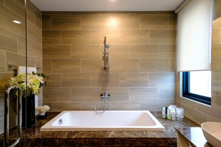 Moderne badkamer met een rechthoekig bad, beige tegelwanden en een douchekop. Een marmeren aanrechtblad bevat toiletartikelen en bloemstukken. Een raam met een jaloezie is aan de rechterkant.