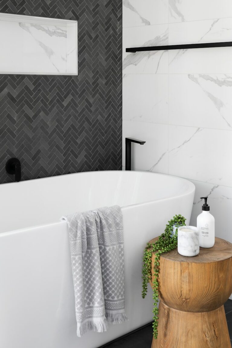 Moderne badkamer met een wit bad, grijze visgraattegels, houten krukje, grijze handdoek, plant en zeepdispenser.