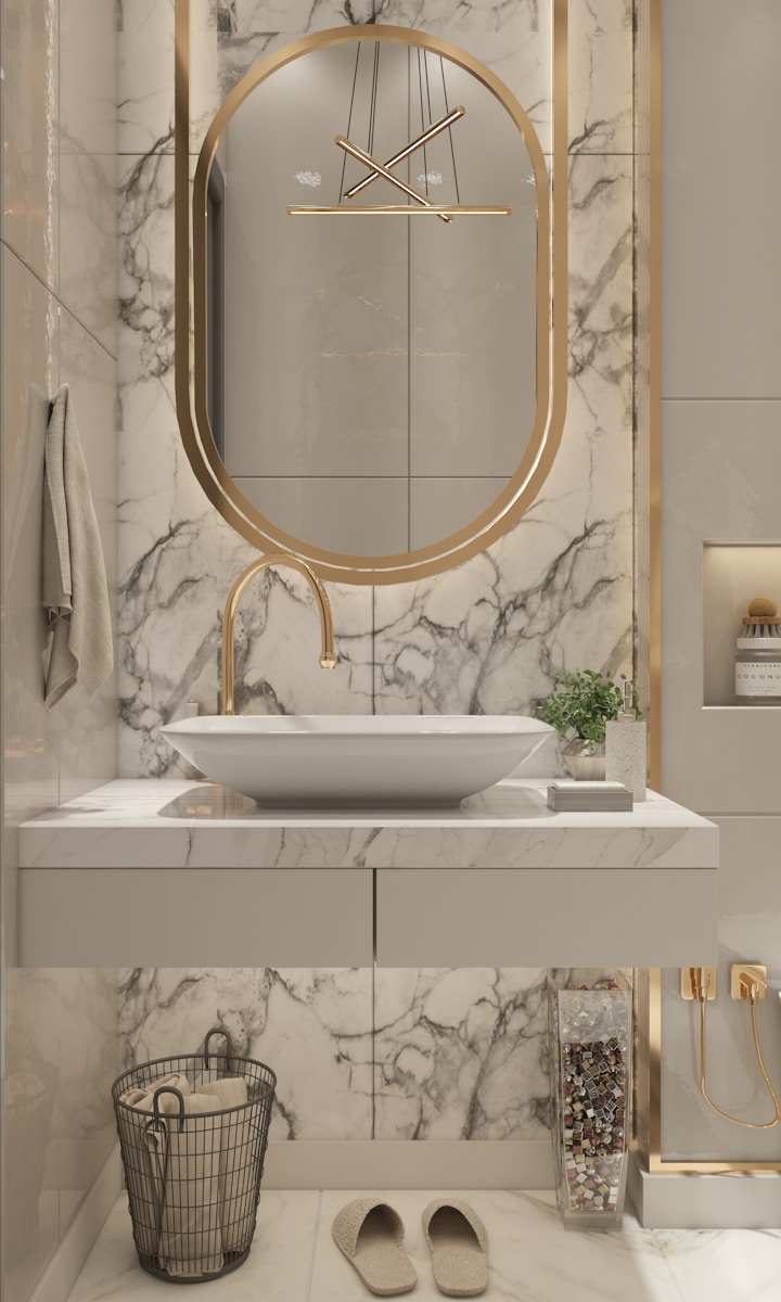 Een moderne badkamer met een marmeren aanrecht, ovale spiegel, gouden accenten en een witte wastafel. Slippers en een handdoek liggen op de vloer. Een kleine plant en diverse toiletartikelen staan op het aanrecht.