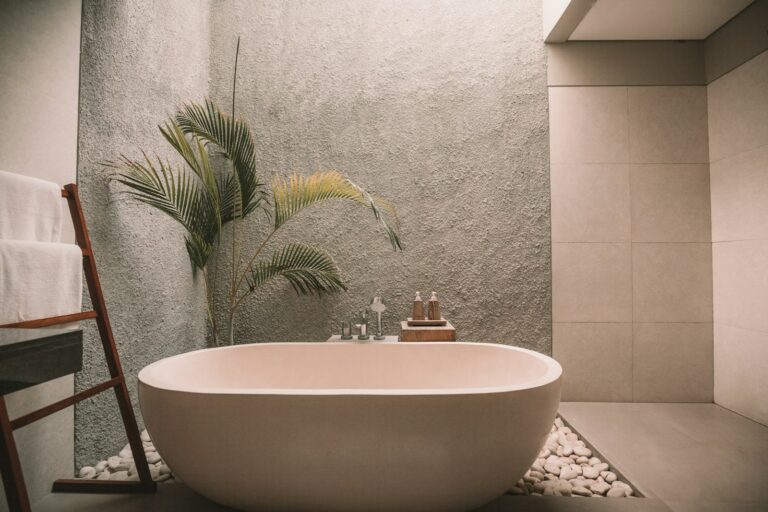 Minimalistische badkamer met een vrijstaand ovaal bad omringd door decoratieve stenen. Een houten rek houdt witte handdoeken vast en een potplant staat bij een getextureerde muur.