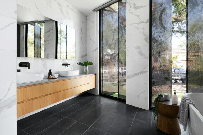Moderne badkamer met twee wastafels op een houten wastafelmeubel, muren met marmerpatroon, grote ramen en een kleine houten kruk naast een vrijstaand bad. Natuurlijk licht filtert door de bomen buiten.