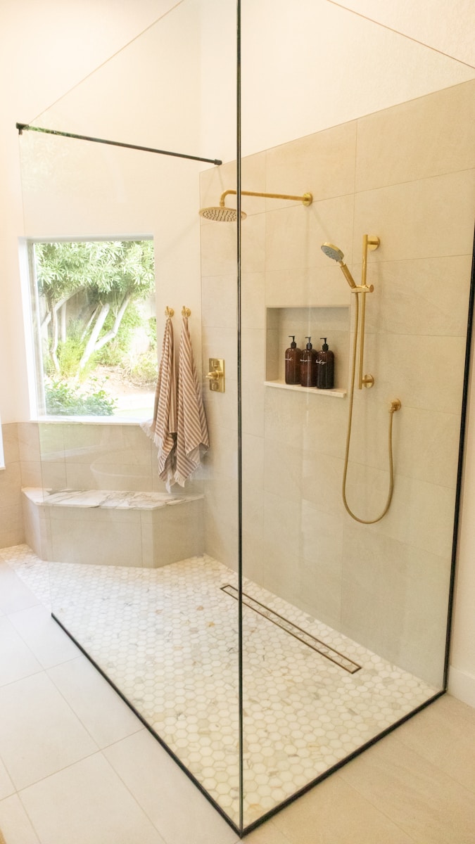 Moderne douche met glazen wanden, gouden armaturen, een ingebouwde plank met flessen en een raam met uitzicht op groen. Gestreepte handdoeken hangen aan de muur bij een klein bankje.