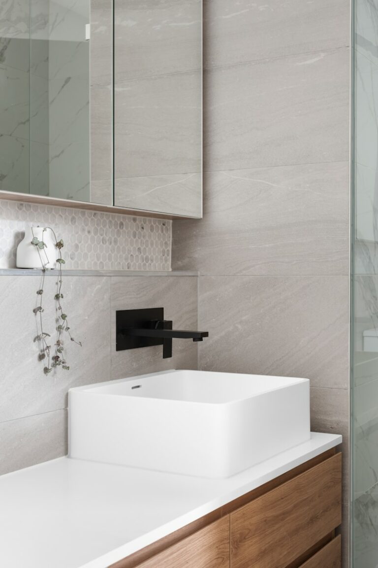 Moderne badkamer met een witte rechthoekige wastafel, zwarte kraan, houten kast en een minimalistische plantendecoratie. Marmeren muren en een glazen douchepaneel zijn zichtbaar.
