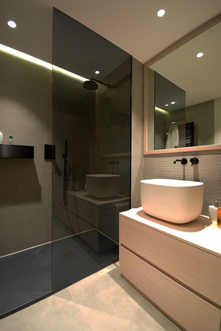 Moderne badkamer met een regendouche met glazen wand, een grote witte wastafel op een houten onderstel, een rechthoekige spiegel en inbouwverlichting.