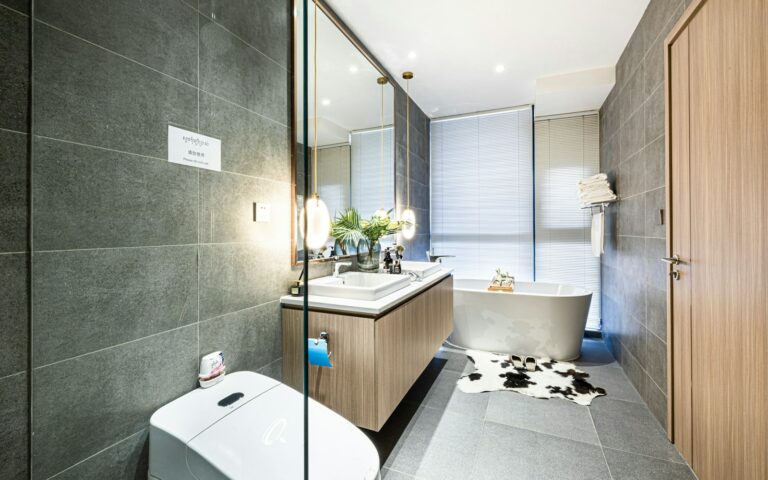 Moderne badkamer met grijze tegels, met een vrijstaand bad, spiegelmeubel met wastafel en wandcloset. Er ligt een koeienhuidkleed op de vloer en planten sieren het aanrecht.