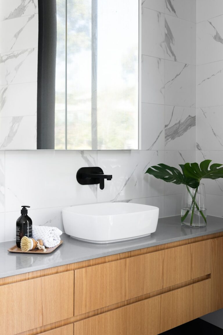 Moderne badkamer met een rechthoekige witte wasbak op een houten kast, zwarte kraan aan de muur, grote spiegel en een vaas met groene bladeren. Bij de wasbak staan een zeepdispenser en toiletartikelen.