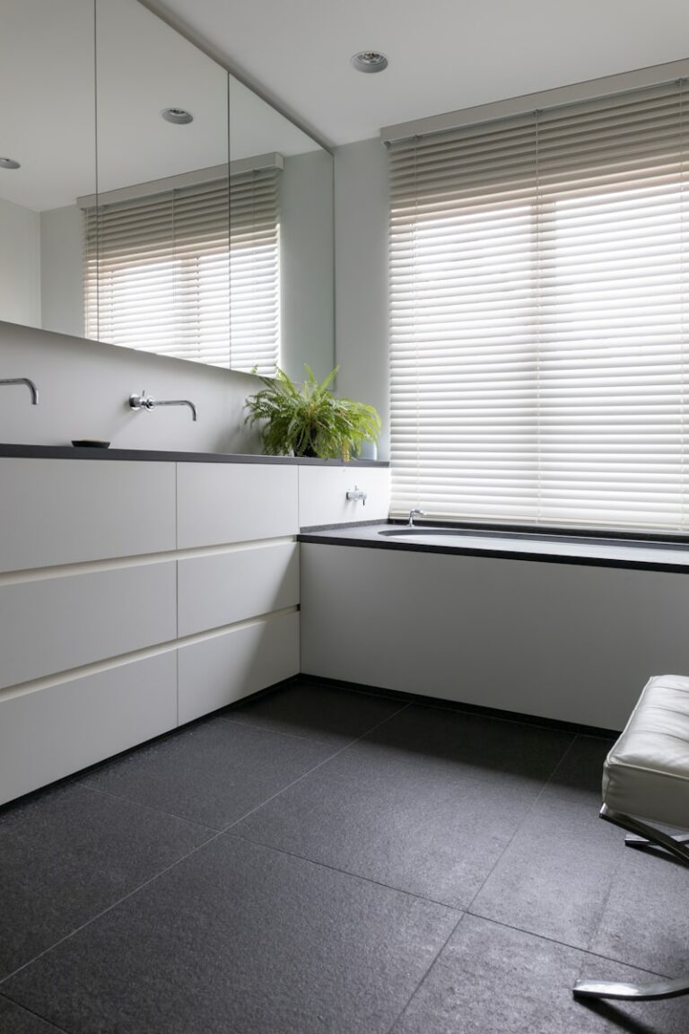 Moderne badkamer met een witte wastafel, spiegelwand en zwart aanrecht. Licht stroomt door grote ramen met beige jaloezieën; kleine plant op het aanrecht. Zwarte betegelde vloer.