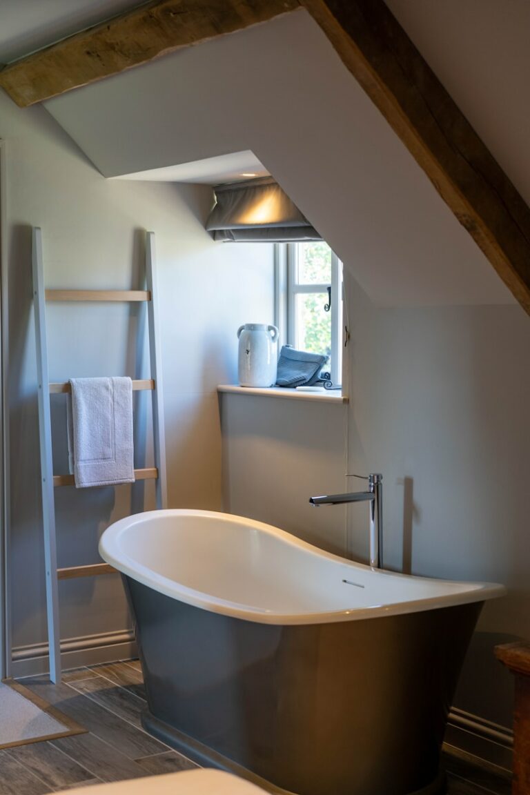 Badkamer met een vrijstaand bad, een ladder-stijl handdoekrek met een handdoek en een klein raam. Houten balken en natuurlijk licht versterken de ruimte.