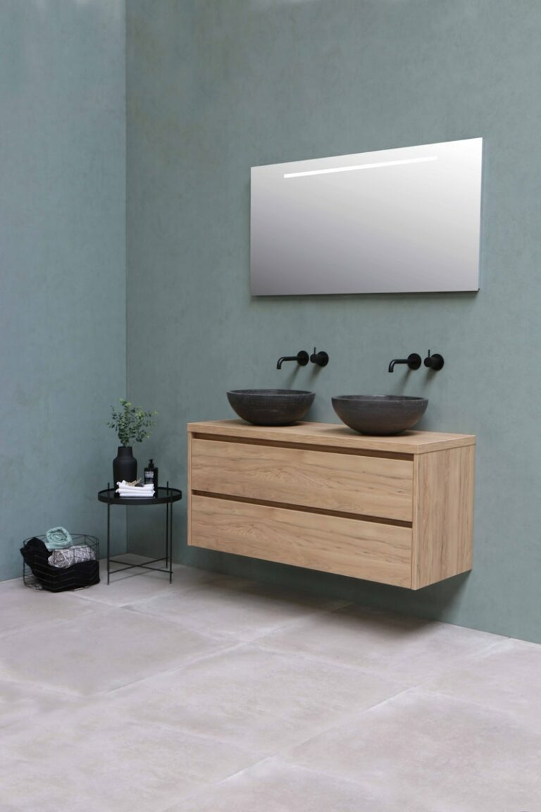 Moderne badkamer met twee donkere ronde wastafels op een zwevend houten meubel, zwarte kranen, een grote rechthoekige spiegel en een bijzettafel met decoratieve elementen tegen een gedempt groene muur.